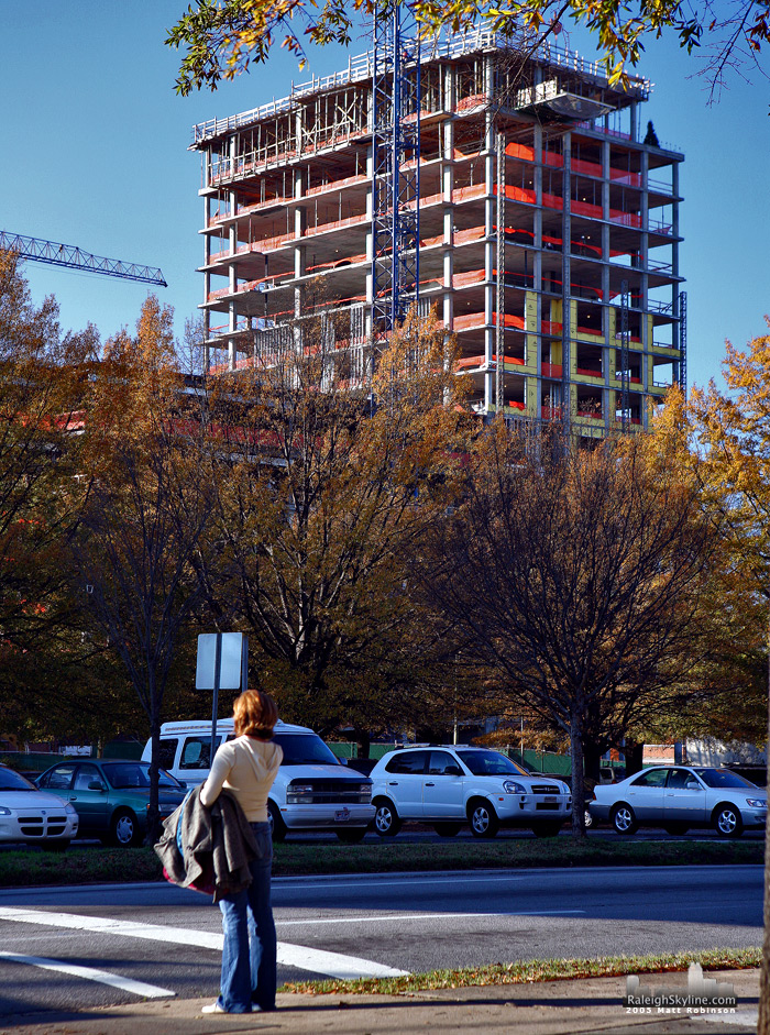 A woman awaits to cross Dawson Street as the Quorum Center peeks over the trees. 