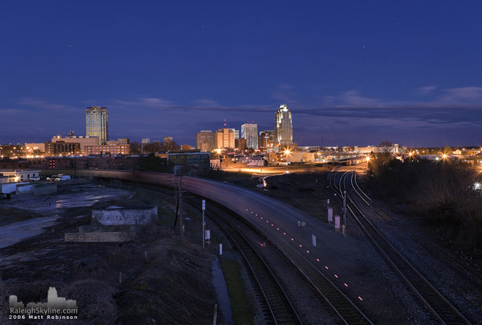 The last car of a long train sweeps past downtown Raleigh.